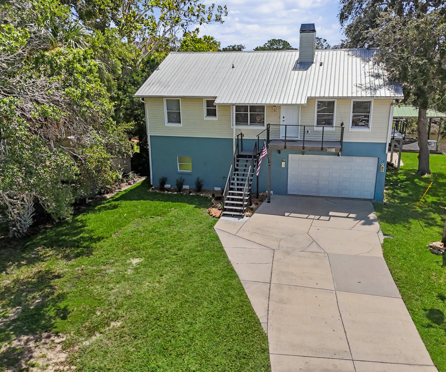 Elevated view of the front of The Rusty Hook Riverhouse — cream siding over a shallow-water blue base, metal roof, stair to the front deck and a long concrete driveway