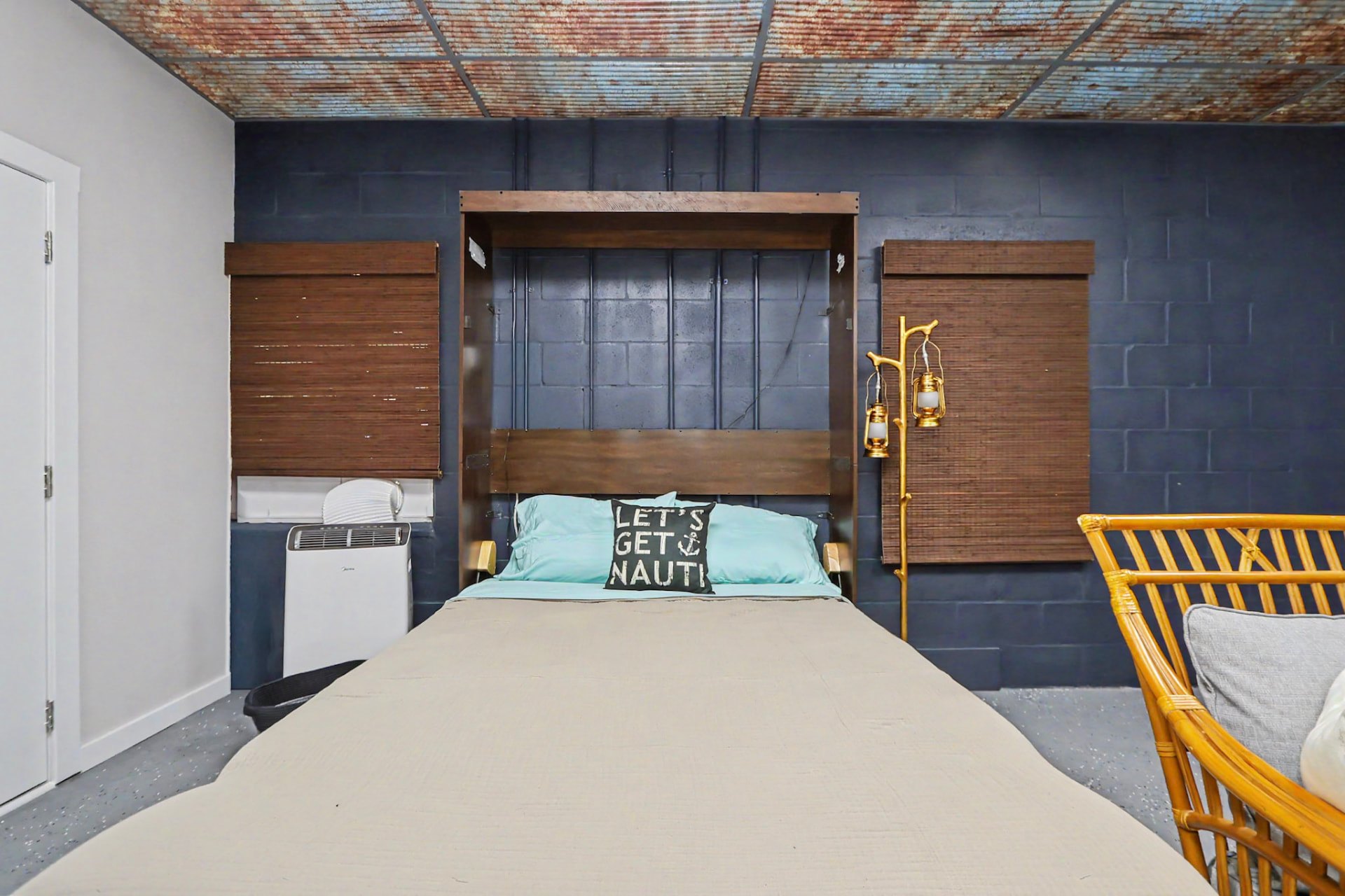 The downstairs Murphy bed room made up for the night — a queen Murphy bed folded down against a navy wall beneath a rusted tin-look ceiling, with lantern sconces and bamboo shades