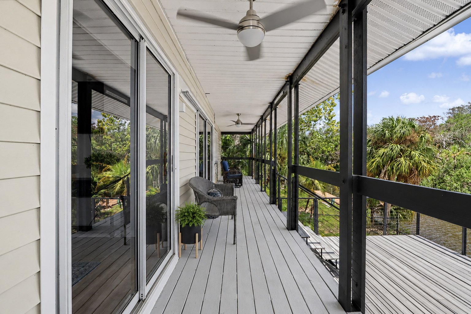 The long open porch running the full length of the house — wicker chairs, a planter, slow ceiling fans and the creek through the cable rails